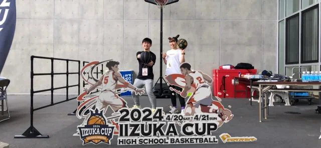 高校バスケットボール大会「IIZUKA CUP 2024」の会場装飾パネル前で記念撮影をする様子。