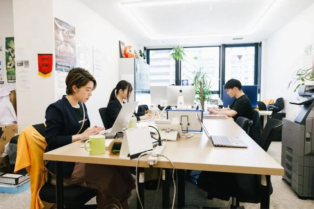 明るいオフィスで複数の社員がパソコンを使用し、それぞれの業務に取り組んでいる様子。