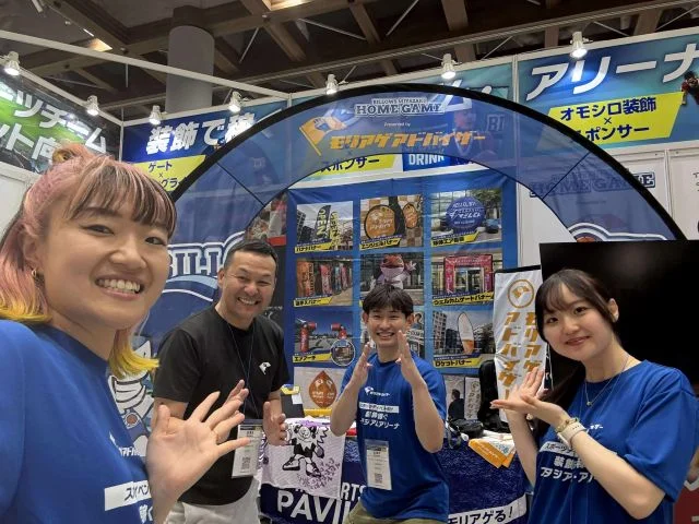 展示会のブース前でスタッフが商品を手に記念撮影する様子。スポーツ関連イベントでの出展・交流風景。